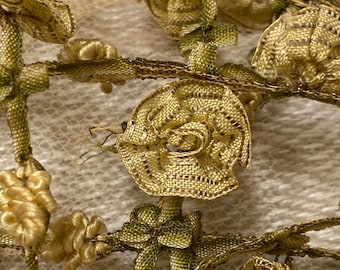 May include: Close-up of a gold and green fabric trim with a floral design. The trim is made of delicate, woven fabric and features small, intricate flowers.