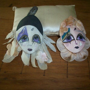 May include: Two handmade fabric brooches with painted faces. One has a black hat and purple and blue accents. The other has a pink face with a tear and a blue and green accent.