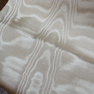 May include: A piece of off-white fabric featuring a fine, horizontal ribbed texture. The material displays a subtle, repeating pattern of vertical bands created by slightly raised, thicker ridges that interrupt the consistent linear weave.