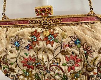May include: Cream-colored vintage handbag with floral embroidery in shades of red, orange, blue, and green. The bag features a gold chain strap and a decorative clasp with red accents. The bag is embellished with small, sparkling rhinestones.