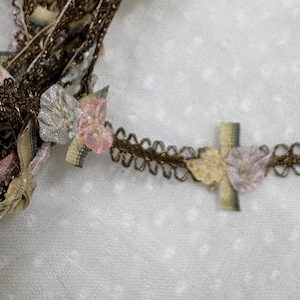 Op de afbeelding: Close-up van decoratieve sierband met een bruine metalen rand en kleine, delicate bloemaccenten in roze, lichtblauw en geel. De sierband is ingewikkeld ontworpen met een geschulpte rand en kleine strikjes. De achtergrond is een witte stof met een subtiel stippenpatroon.
