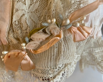May include: Close-up of a delicate white lace fabric with a pink satin ribbon and pearl embellishments. The lace has a floral pattern and is trimmed with a scalloped edge.