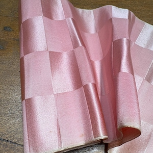 May include: A roll of light pink fabric featuring a repeating checkered pattern with a subtle sheen. The material is folded in soft waves, displaying a smooth, satin-like texture across the square blocks that alternate in light and dark pink tones.