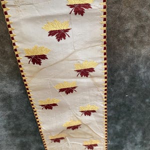 Può includere: Tessuto color crema con un motivo ripetuto di foglie ricamate in oro e bordeaux. Il bordo è rifinito con un bordo bordeaux e cuciture dorate. Il tessuto sembra vintage.