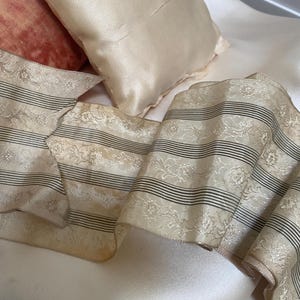 Könnte beinhalten: Nahaufnahme eines cremefarbenen Bandes mit schwarzen horizontalen Streifen und floralen Mustern. Das Band ist über einer Satin-Oberfläche drapiert, mit zwei kleinen Satin-Kissen im Hintergrund. Die Kissen sind hellpfirsichfarben.