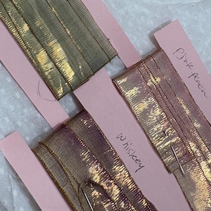May include: A close-up of three samples of metallic ribbon in gold, pink, and bronze. Each ribbon is attached to a pink card with handwritten labels: "Blue", "Pink peach", and "Whiskey".