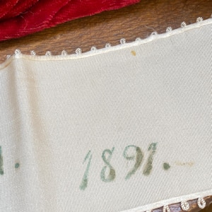 Może przedstawiać: Zbliżenie kremowej wstążki z numerem "1891" wydrukowanym zielonym tuszem. Wstążka ma dekoracyjne obramowanie i jest umieszczona na drewnianej powierzchni. Czerwona i czarna tkanina widoczne w tle.