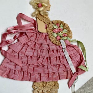May include: A vintage-style paper doll with a blonde wig and a pink ribbon dress. The doll holds a bouquet of pink and green flowers. The dress features layered ribbons, and the doll wears silver shoes. The overall design is reminiscent of a bygone era.