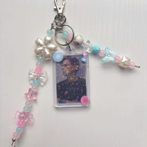 Op de afbeelding: Een sleutelhanger met een rechthoekige doorzichtige plastic fotohouder met een portret van een persoon, versierd met twee kleine bloemstickers. Bevestigd zijn twee kralenstrengen met roze, blauwe en witte kralen, een grote bloem en een vlinder.