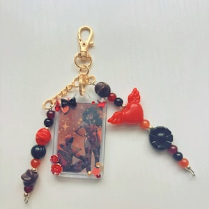 May include: A rectangular keychain featuring a clear plastic frame containing an illustration of two figures, accented with a gold-tone lobster clasp, black and red beads, small red rose charms, a red winged heart charm, and a black flower bead.