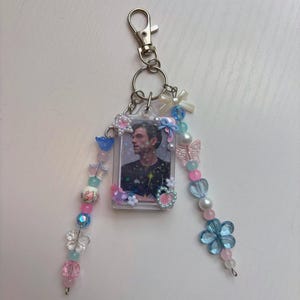 May include: A rectangular photo holder keyring featuring a portrait of a person in a black shirt. The frame is decorated with small charms including stars, hearts, and bows. Two beaded strands hang from the frame, featuring blue, pink, and white beads in various shapes like flowers, butterflies, and spheres.