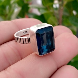 May include: A silver ring featuring a large, rectangular, faceted dark blue gemstone set in a smooth metal bezel. The band has a textured, vertical-line pattern along the outer surface and is stamped with the number 925 on the inner side.