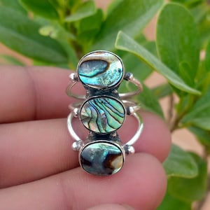 Könnte beinhalten: Ein Silberring mit drei vertikal übereinander angeordneten ovalen Abalone-Muschel-Cabochons. Die irisierende Muschel zeigt wirbelnde Muster in Grün-, Blau- und Brauntönen. Die Metallschiene ist in drei separate Abschnitte unterteilt.