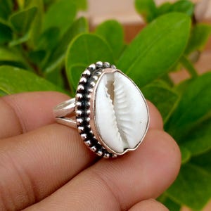 Peut inclure: Une bague en argent avec un coquillage cauri blanc au centre. Le coquillage est enchâssé dans une monture en argent décorative avec un bord perlé. L'anneau de la bague est en argent poli et présente un design fendu sur les côtés.