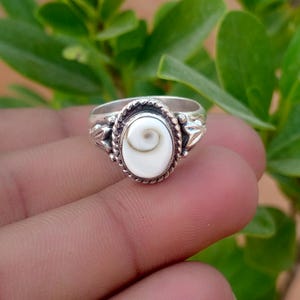Puede incluir: Un anillo de plata con una incrustación ovalada de concha de ojo de shiva blanca en el centro. La banda de metal está decorada con un borde trenzado tipo cuerda que rodea la concha y grabados detallados en forma de hoja en los laterales.