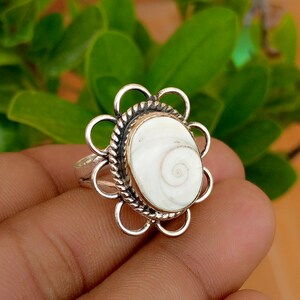Puede incluir: Un anillo de plata con una piedra central ovalada de concha de ojo de Shiva blanca. La piedra está rodeada por un borde de metal trenzado y un marco exterior de ocho bucles circulares de plata, creando un diseño en forma de flor.