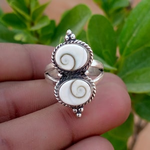 Puede incluir: Un anillo de plata con dos incrustaciones ovaladas de concha de ojo de Shiva blanca, cada una con un patrón de espiral marrón natural. El engaste está enmarcado por un borde de metal trenzado y pequeñas cuentas de plata en los extremos.
