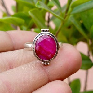 May include: A silver ring featuring a large, oval-shaped, faceted hot pink gemstone set in a smooth metal bezel. The ring band is plain silver, and the setting is accented with small, decorative silver beads at the top and bottom of the stone.