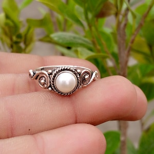 Puede incluir: Un anillo de plata con una perla blanca redonda central engastada en un borde de cuerda trenzada. La banda muestra intrincados adornos de metal en forma de volutas y espirales a ambos lados del engaste de la perla, con un acabado pulido.