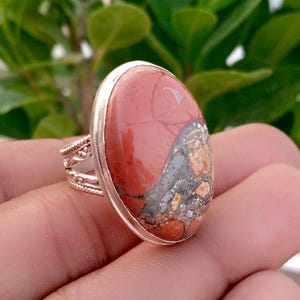May include: A silver ring featuring a large, oval-shaped cabochon stone with a smooth, polished surface. The stone displays a mix of terracotta red, grey, and beige mottled patterns. The silver band has an intricate, decorative filigree design.