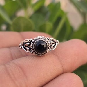 May include: A silver ring featuring a central round black stone set within a twisted rope-style border. The band displays intricate metalwork with decorative scroll patterns and small silver beads on either side of the main setting.