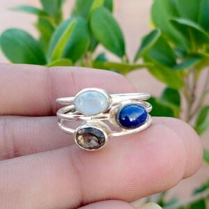 Peut inclure: Une pile de trois bagues en argent, chacune ornée d'une pierre précieuse ovale unique. La bague supérieure porte une pierre blanche translucide, celle du milieu une pierre bleu profond et l'inférieure une pierre brune à facettes.