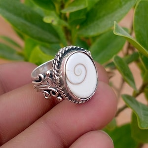 May include: A silver ring featuring an oval-shaped white shiva eye shell inlay with a natural spiral pattern. The metal band is decorated with a twisted rope border and intricate scrollwork detailing on the sides of the setting.