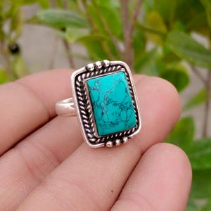 May include: A rectangular silver ring featuring a vibrant turquoise stone with dark veining. The stone is set within a twisted rope-style metal border, accented by small silver spheres at the top and bottom edges of the rectangular setting.