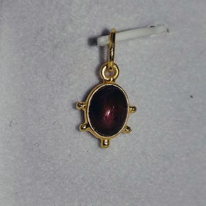 Può includere: Un ciondolo color oro con una pietra cabochon ovale di colore viola scuro, incastonata in una montatura metallica con quattro piccole punte decorative che sporgono dai lati. Il ciondolo presenta un piccolo anello circolare sulla parte superiore.