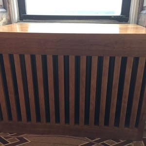 Puede incluir: Una cubierta de radiador de madera con acabado marrón claro. La parte frontal presenta listones verticales, y la parte superior es una superficie sólida y plana. La cubierta está colocada frente a una ventana, lo que sugiere su uso decorativo y funcional.