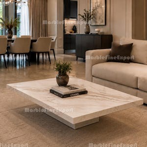 Puede incluir: Una mesa de centro rectangular con una superficie de mármol blanco con vetas grises y una base sólida. Un pequeño jarrón oscuro con vegetación y una pila de dos libros, incluyendo uno titulado CHANEL, se encuentran en el centro de la mesa.