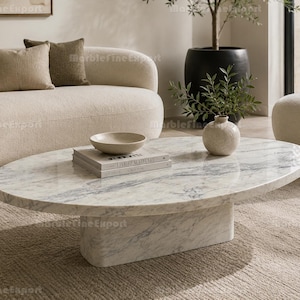 May include: An oval coffee table crafted from white marble with grey veining, featuring a solid, thick pedestal base. The tabletop holds a stack of two books, a shallow beige bowl, and a small, textured, round off-white ceramic vase.