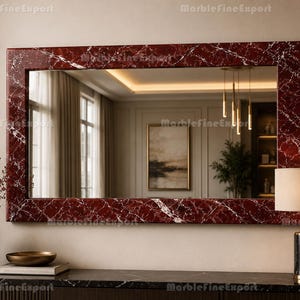 May include: A rectangular wall mirror featuring a deep red marble frame with prominent white veining. The mirror reflects a room interior with a framed picture, hanging pendant lights, and a plant. The text MarbleFineExport is visible.