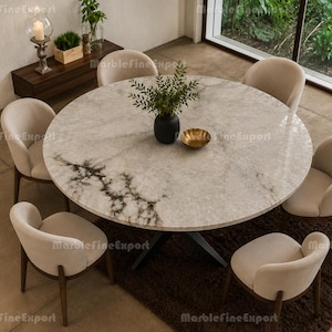 Op de afbeelding: Een ronde eettafel met een licht marmeren blad met donkere aders, ondersteund door een donker metalen onderstel. Zes gestoffeerde stoelen met gebogen rugleuningen en houten poten staan rond de tafel. Een zwarte vaas met groen staat in het midden.