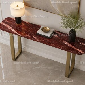 Puede incluir: Una mesa consola larga con un tablero de mármol rojo oscuro pulido con vetas blancas y dos patas de metal dorado cepillado en forma de U. La superficie de la mesa sostiene una lámpara blanca con base negra, dos libros apilados, un cuenco dorado y un jarrón.