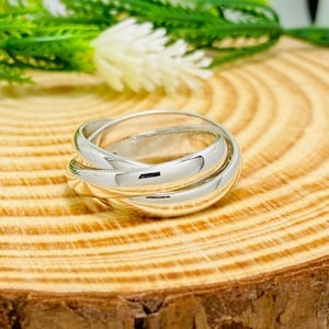 Può includere: Un anello color argento lucido composto da tre fasce intrecciate che si sovrappongono per formare un unico pezzo continuo. La superficie metallica è liscia e riflettente, mostrando un profilo arrotondato che crea un aspetto a strati.