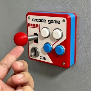 Op de afbeelding: Een aan de muur gemonteerde arcade game controller met een rode joystick, twee witte knoppen en twee blauwe knoppen. De witte voorplaat toont de tekst arcade game en coin met een zilveren muntgleuf. De unit heeft een rood en blauw frame.
