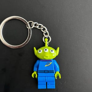 Könnte beinhalten: Ein Schlüsselanhänger mit einer kleinen, lindgrünen, dreiäugigen Alien-Figur mit spitzen Ohren. Die Figur trägt eine blaue Uniform mit violettem Kragen und einem gelben Planeten-Emblem auf der Brust. Sie ist an einem runden silbernen Metallring befestigt.
