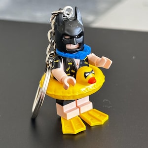 Pode incluir: Um porta-chaves de minifigura do Batman com capuz preto, colete preto com símbolos de morcego amarelos e um tubo de respiração azul ao pescoço. A figura usa uma boia insuflável de pato amarelo brilhante e barbatanas amarelas nos pés.