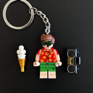 May include: A keyring featuring a plastic minifigure with brown hair, green goggles, a red shirt with a yellow duck pattern, and green shorts. Beside the figure are a white ice cream cone accessory and a black portable radio accessory.