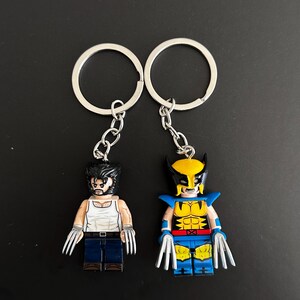 Peut inclure: Deux porte-clés en métal représentant des figurines miniatures d'un personnage aux cheveux noirs et aux favoris. L'une porte un débardeur blanc et un pantalon bleu, l'autre un costume jaune et bleu avec un masque. Les deux ont des griffes argentées.