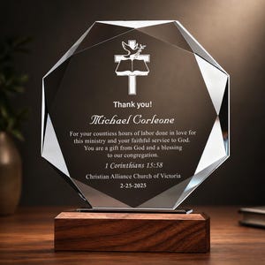 May include: An octagonal glass award plaque featuring a white engraved cross and dove icon above the text Thank you! Michael Corleone, followed by a message of appreciation, 1 Corinthians 15:58, the church name, and the date 25-02-2025 on a wooden base.
