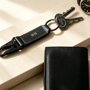 May include: A black leather keychain featuring white stitching and the gold embossed initials WH. The keychain includes a black metal carabiner clip and a silver-toned metal key ring holding two Yale keys, positioned near a black leather wallet.