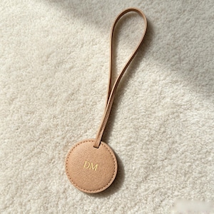 May include: A circular beige leather luggage tag featuring a textured surface and a long, thin matching strap. The centre of the circular tag is embossed with the gold-coloured capital letters DM, providing a clear and personalised detail.