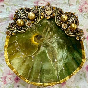May include: A decorative shell dish featuring a central image of a winged fairy in a green forest setting. The top edge is adorned with ornate gold-toned metalwork, large gold pearls, clear rhinestones, and a small central cameo portrait.