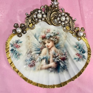 May include: A decorative scallop shell featuring a central illustration of a figure with floral headwear and wings. The shell is framed by an ornate gold-toned metal border adorned with white faux pearls and small clear crystals along the top edge.