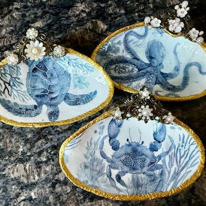 Op de afbeelding: Drie decoratieve schelpschalen met blauw-witte illustraties van een zeeschildpad, een octopus en een krab. Elke schelp heeft een goudkleurige rand en is versierd met een bronskleurige metalen decoratie met kleine witte bloemetjes.