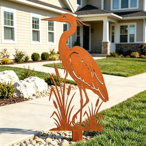 Könnte beinhalten: Ein rostfarbener Gartenstecker aus Metall mit der Silhouette eines Reihers inmitten von Rohrkolben. Das flache, ausgeschnittene Design zeigt den Vogel im Profil mit langem Hals und spitzem Schnabel, umgeben von hohen, schlanken Schilfhalmen.