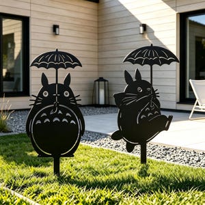 Può includere: Due picchetti da giardino in metallo nero con sagome di una creatura rotonda con orecchie che tiene un ombrello. Una figura è in piedi con una pancia a motivi, mentre l'altra è in una posa dinamica, entrambe montate su sottili aste metalliche.