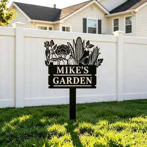 Pode incluir: Uma estaca de jardim de metal preto com uma silhueta de vários vegetais, incluindo milho, brócolis e cenouras, acima de uma placa retangular com o texto MIKE'S GARDEN em letras maiúsculas brancas, montada em um poste vertical.